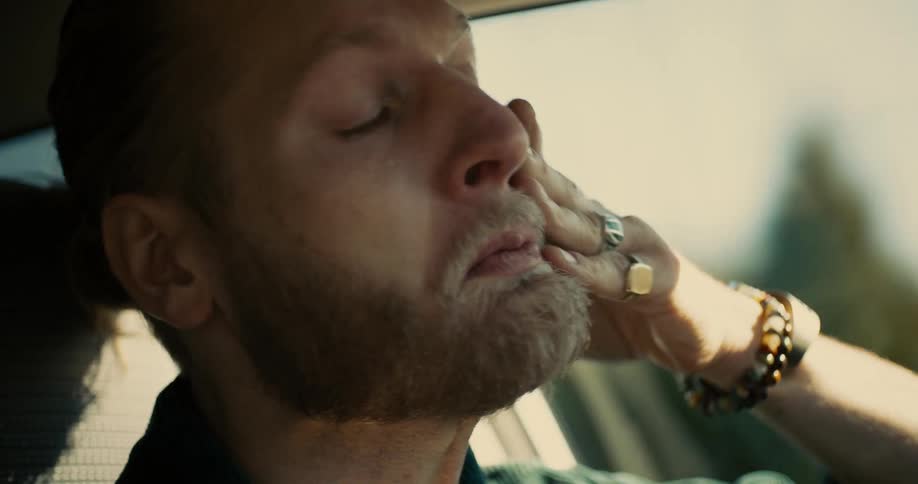 A young man with a ponytail and a full beard drives a car at midday tears streaming down his face in sadness