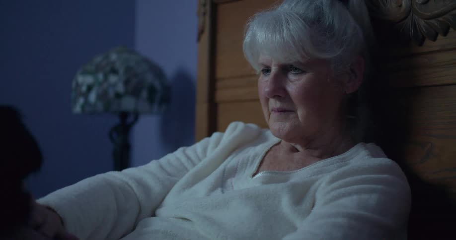An elderly woman gently pets her dog while sitting in a bedroom