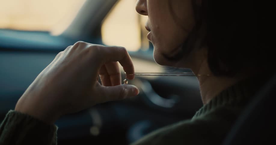 A young woman sits in the passenger seat of a car parked by the side of the road playing with her necklace