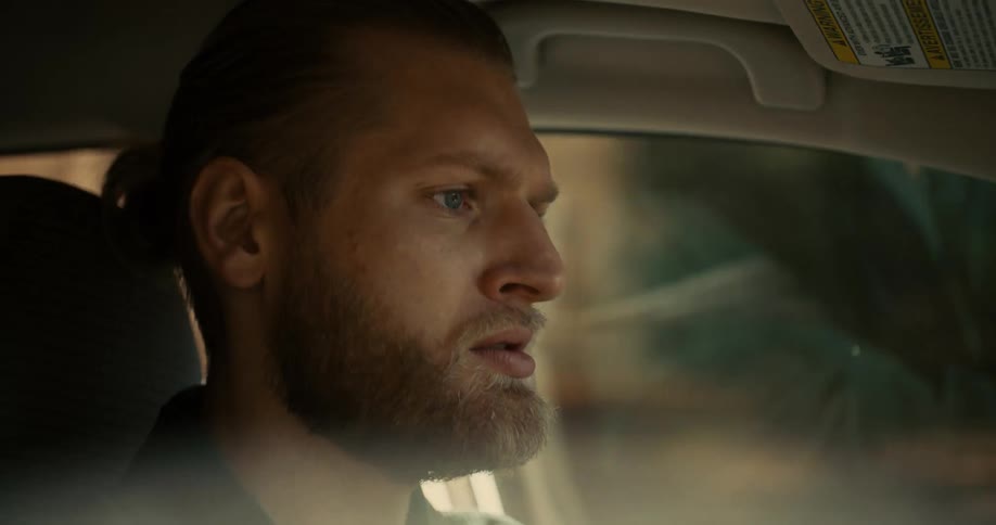 A man with light colored eyes and a brown beard is shown in a close side view from the passenger seat looking straight ahead as he sits in the drivers seat of a car