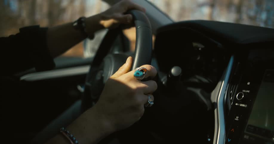 A driver with a large turquoise and coral ring on one hand and a black bracelet on the other keeps both hands on a cars steering wheel while driving