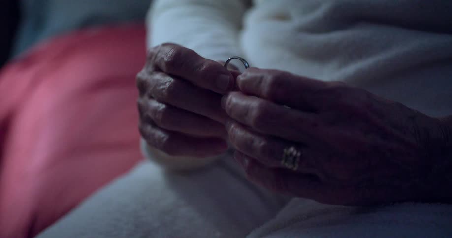 A pair of wrinkled hands belonging to an elderly person is shown in a closeup shot gently holding and turning a simple silver ring the hands and ring are in focus while the background is softly blurred