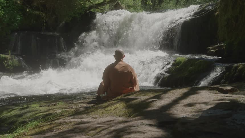 Monk sitting on rocks at the edge of a waterfall in nature