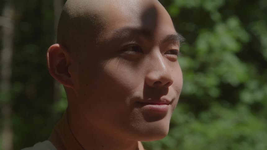 Based on the images from the video a closeup shot of a young man with a shaved head is shown he is looking off to the side and smiling slightly with dappled sunlight visible on his face and shoulders and a blurred background of green foliage