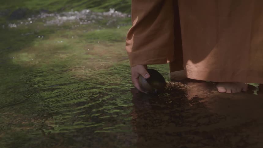 A person in a robe stands barefoot in shallow water while holding a small bowl