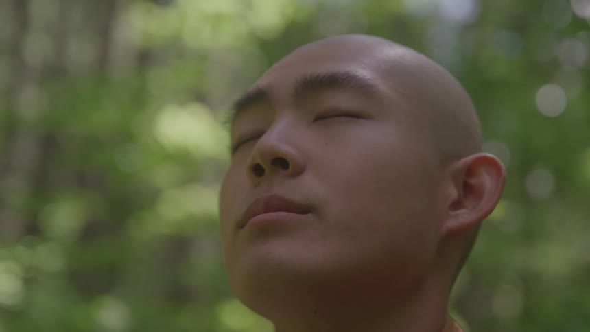 The video shows a closeup of a person with a shaved head standing in a forest with their eyes closed and their face tilted slightly upward the person appears to be meditating or enjoying the peaceful surroundings