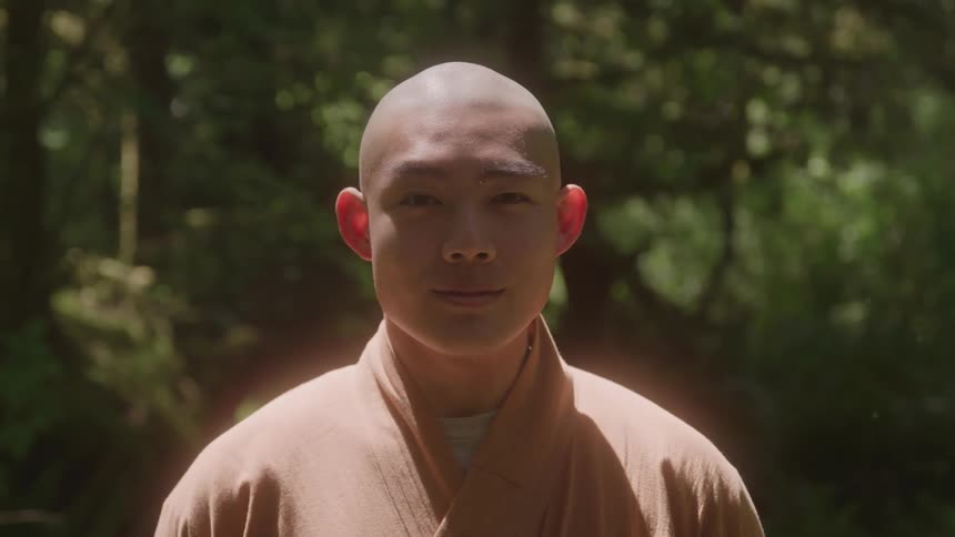 A young tibetan buddhist monk with a shaved head and a serene expression is smiling while standing in a sun dappled forest wearing a light orange robe over a white collared shirt