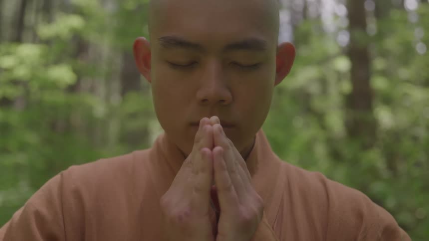 In a serene closeup a young monk with his eyes closed holds his hands together in a prayer position while meditating in a quiet forest