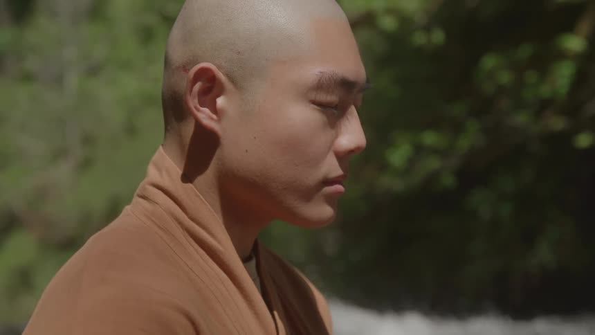 A bald man with his eyes closed in a state of meditation in a peaceful natural outdoor setting