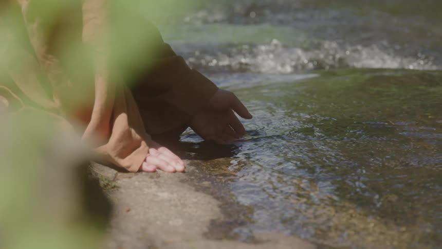 Shot from behind a plant a monk wearing robes kneels at the edge of a rocky stream and gently places his hand into the clear flowing water