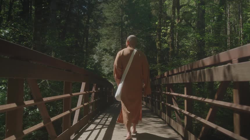 A person wearing a brown robe and carrying a white bag walks barefoot across a wooden bridge through a lush green forest