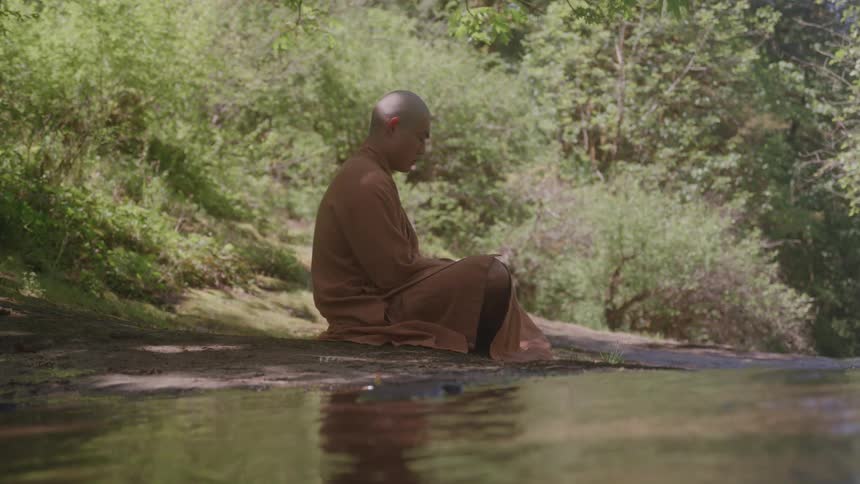 A bald tibetan monk wearing a brown robe sits calmly on a rocky ledge next to a small stream in a forest