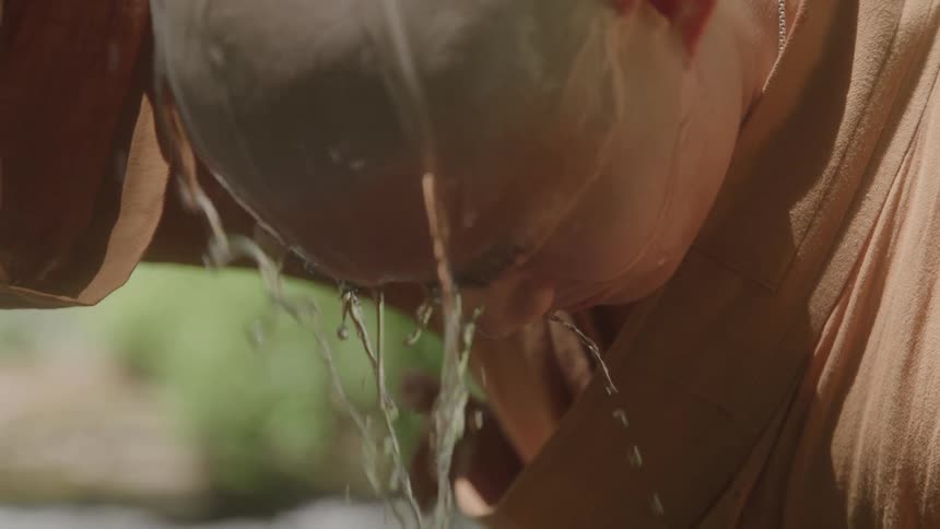 In an intimate slowmotion closeup a monk pours cool water over his shaved head letting it cascade down his face in a cleansing ritual