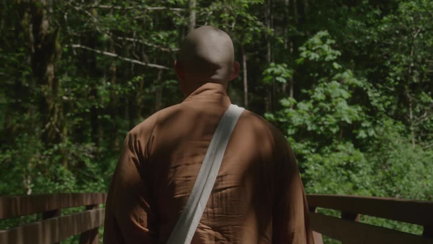 Person in a brown robe crossing a wooden bridge in the forest