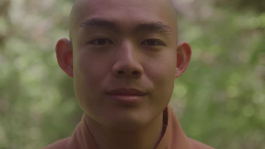 Based on the images from the video the scene is a closeup portrait of a young man with a shaved head he is looking directly at the camera with a neutral and calm expression and the background consists of blurred green foliage