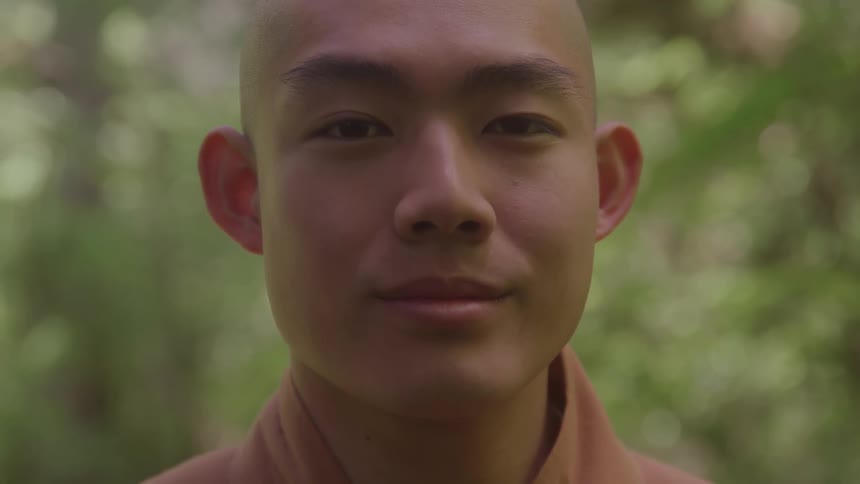 A young bald monk smiles while sitting in the woods at midday