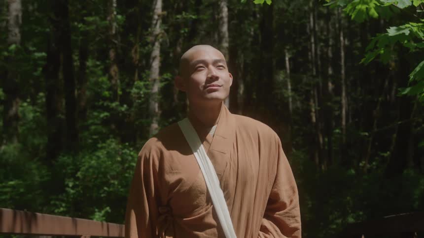 The camera tracks a monk from the waist up as he walks across a bridge in the forest looking up peacefully at the sunlight filtering through the trees