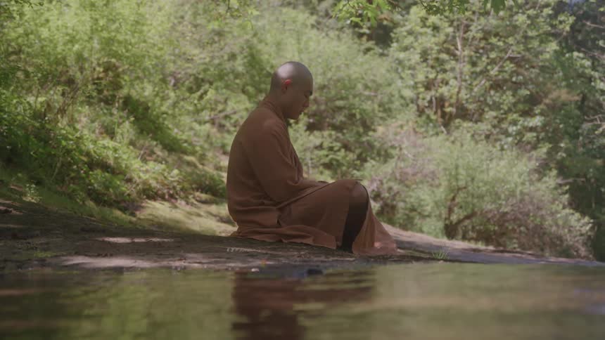 A monk is meditating peacefully by a river in a forest