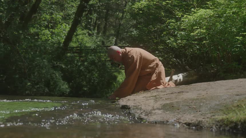 A person in a robe kneeling beside a stream with their hand gently touching the water