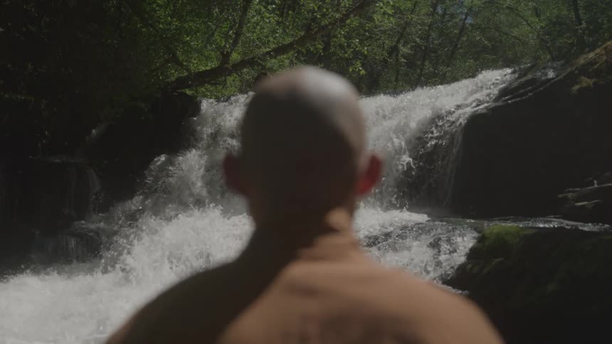 The video shows the back of a persons shaved head as they face away from the camera the person is standing in a lush green forest with a bright flowing waterfall in the background