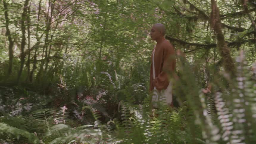 A monk is seen walking through a lush green forest he is wearing a long brown robe and carrying a white bag