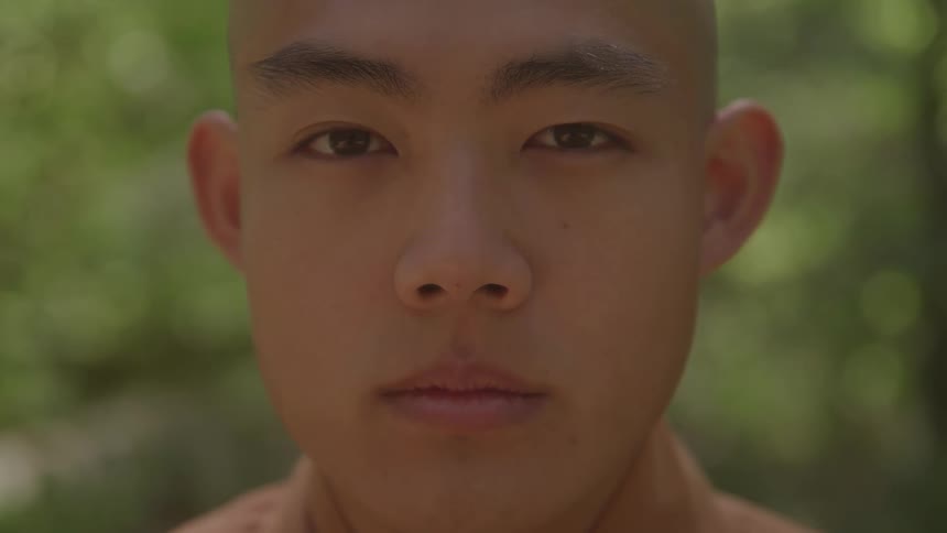 A person with a shaved head and wearing a brown robe initially with their eyes closed then opening them to look directly ahead