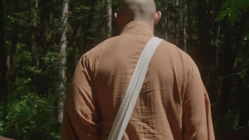The camera starts on the bare feet of a monk walking across a wooden bridge then tilts up his body and continues moving upward into the sunlit leaves of the trees