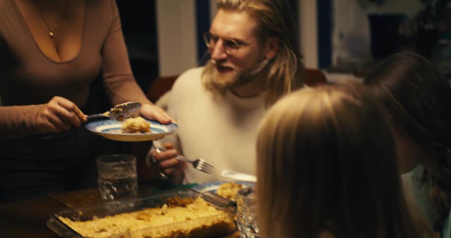 A family gathered at the dining table with a woman serving food from the table onto her plate with a spoon while the others eat