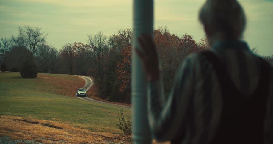 An elderly woman standing waiting for a car that is coming toward her