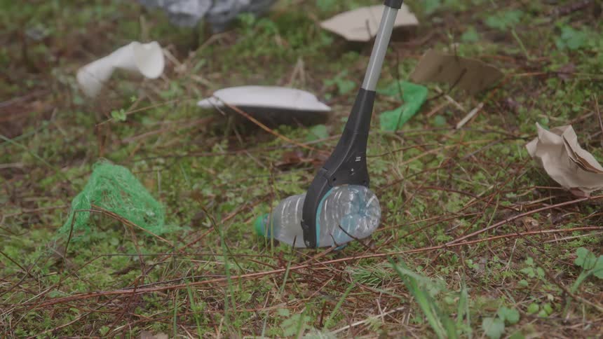 Litter picker grabs a plastic bottle from the ground covered with trash among vegetation