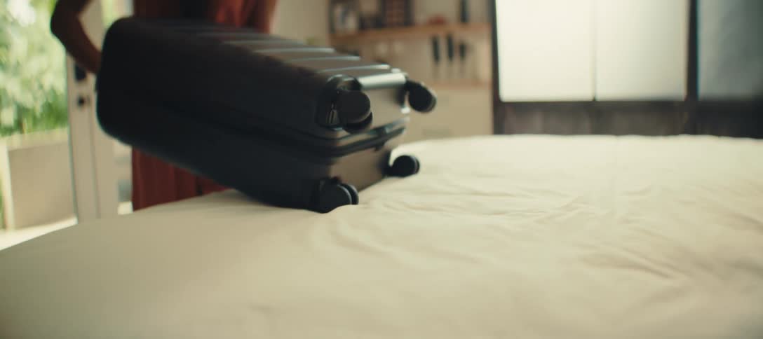 A suitcase is being pulled off a bed and then placed on the floor the suitcase is black and has four wheels