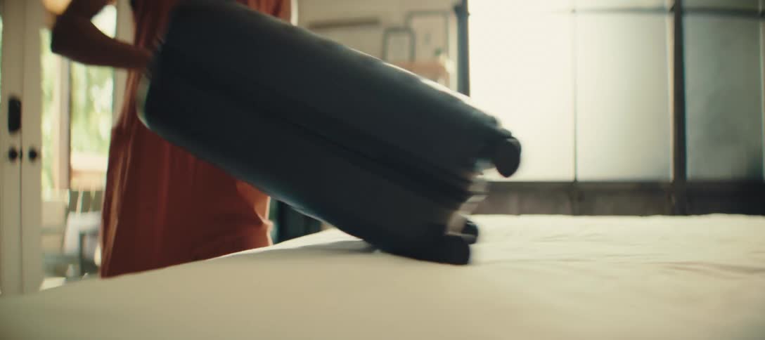 The video shows a person in an orange outfit placing a black suitcase on a bed the person then closes the suitcase and the camera follows the suitcase as they pick it up and walk out of the frame