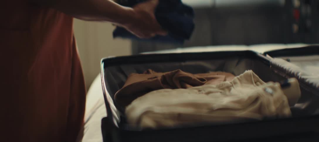 A woman wearing a gold bracelet on their wrist opens a black suitcase and places a blue shirt on top of other folded clothes inside