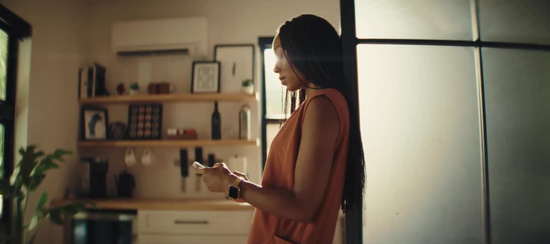 The video shows a young woman standing in a modern kitchen or living room she is looking at her phone which is held in both hands with a focused expression
