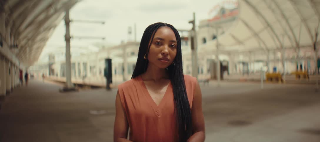 A woman with dark hair styled in long braids stands still and looks directly at the camera while standing under the arched covered canopy of a station