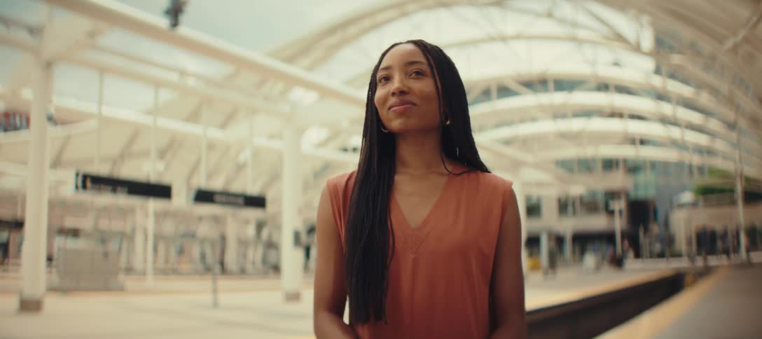 The video shows a young woman with a long braided hairstyle and an orange top she is smiling and looking to her left while standing on what appears to be a train platform