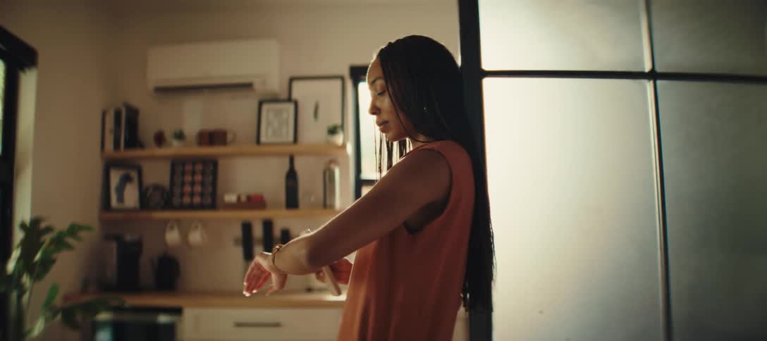 Based on the video a woman with long dark braids and an orange sleeveless shirt is standing in what appears to be a kitchen she is looking at her wrist possibly at a watch or fitness tracker