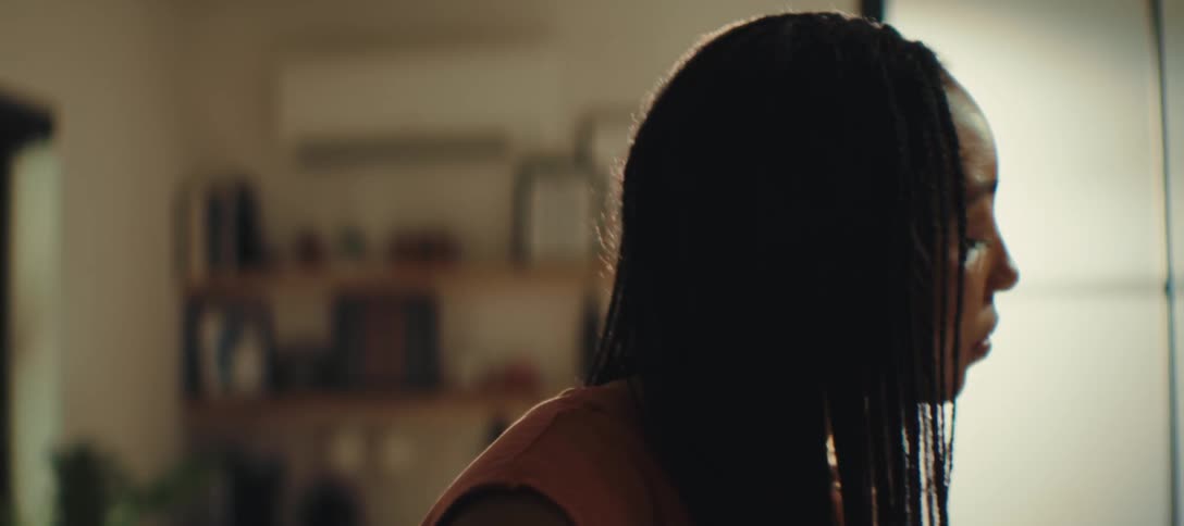 A close side view shows a woman with long dark braids and a rust colored top standing in a sunlit room and looking down with a neutral expression as she moves her head slightly
