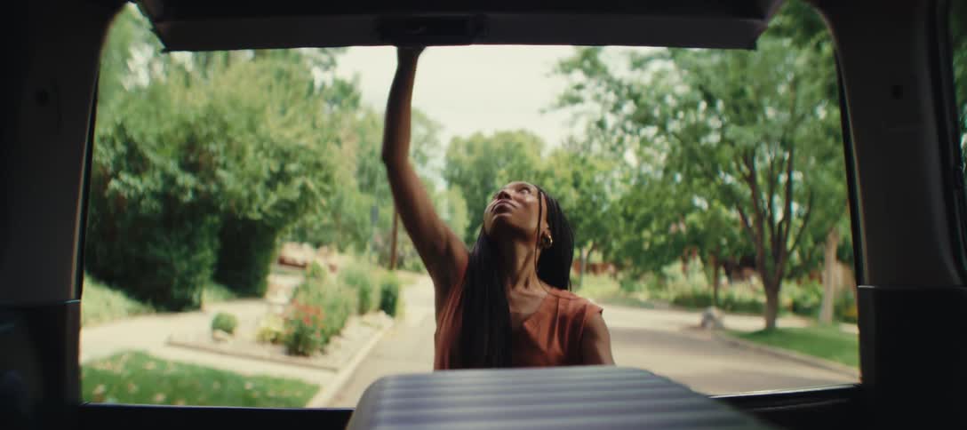 A woman viewed from inside a vehicle putting a suitcase into the car trunk and then closing it