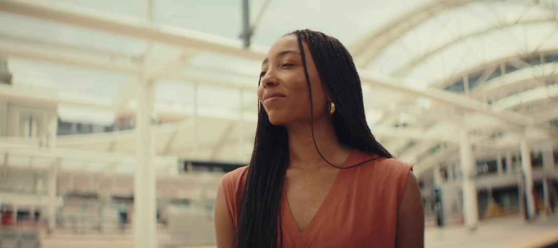 The video shows a woman standing with a black suitcase on a train platform she checks her wristwatch then looks up and smiles thoughtfully into the distance
