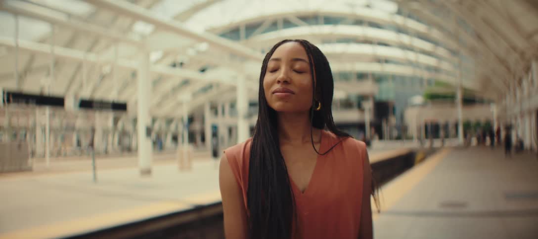 Based on the images from the video a woman with long dark braids is standing on a train platform she is wearing an orange sleeveless top and a gold necklace she stands calmly closes her eyes for a moment and then opens them while looking into the di
