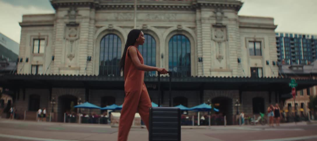 A young woman with long dark hair wearing an orange jumpsuit and white sneakers walks confidently while pulling a black suitcase past a large ornate building with a sign that reads union station