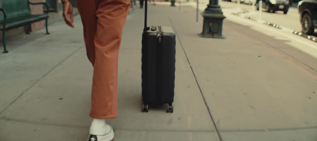 A person wearing rust colored pants and white shoes is walking down a city sidewalk pulling a black four wheeled suitcase behind them