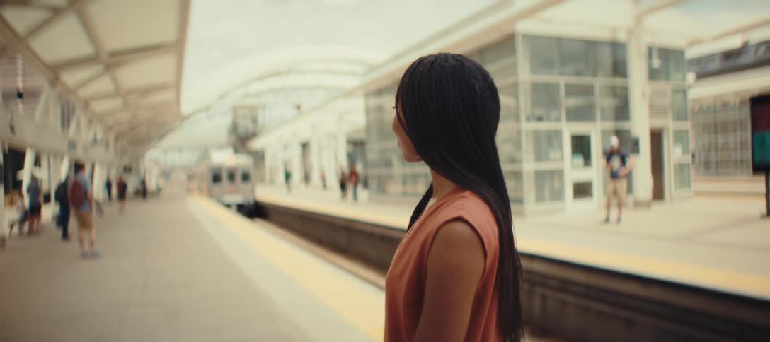 A young woman with braided hair stands in a terminal watching a train arrive at midday