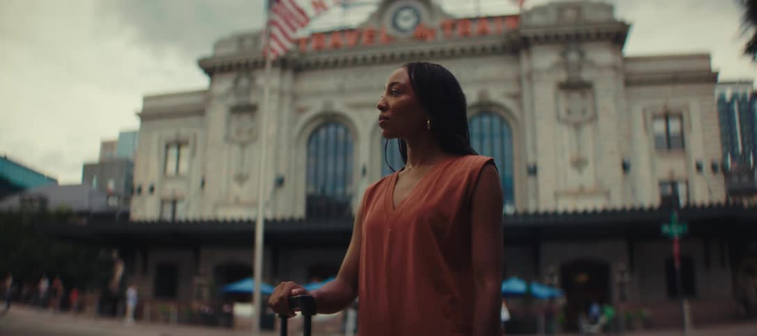 A woman with dark skin and long hair stands in front of a large brick building with a united states flag on top holding the handle of a suitcase and looking away from the camera