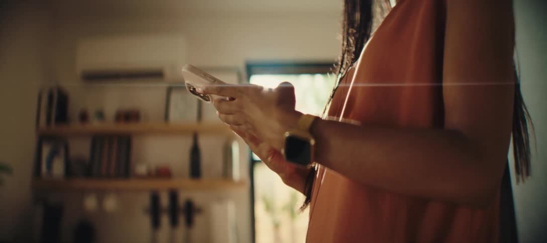 The video shows a closeup of a person holding a smartphone the person is wearing an orange sleeveless top and a gold wristwatch on their left wrist a blurry bookshelf and a window are visible in the background