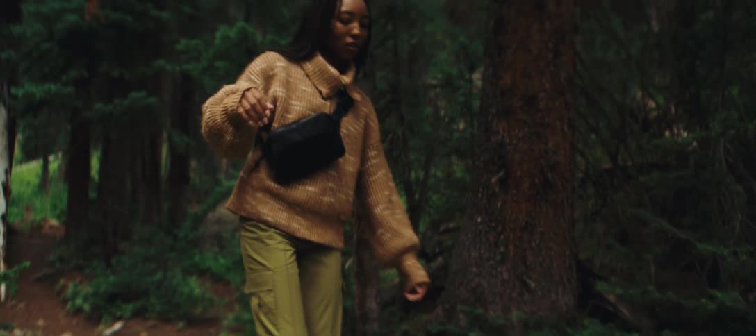 A woman wearing a brown sweater and green pants walks forward while balancing on a fallen log on a forest floor