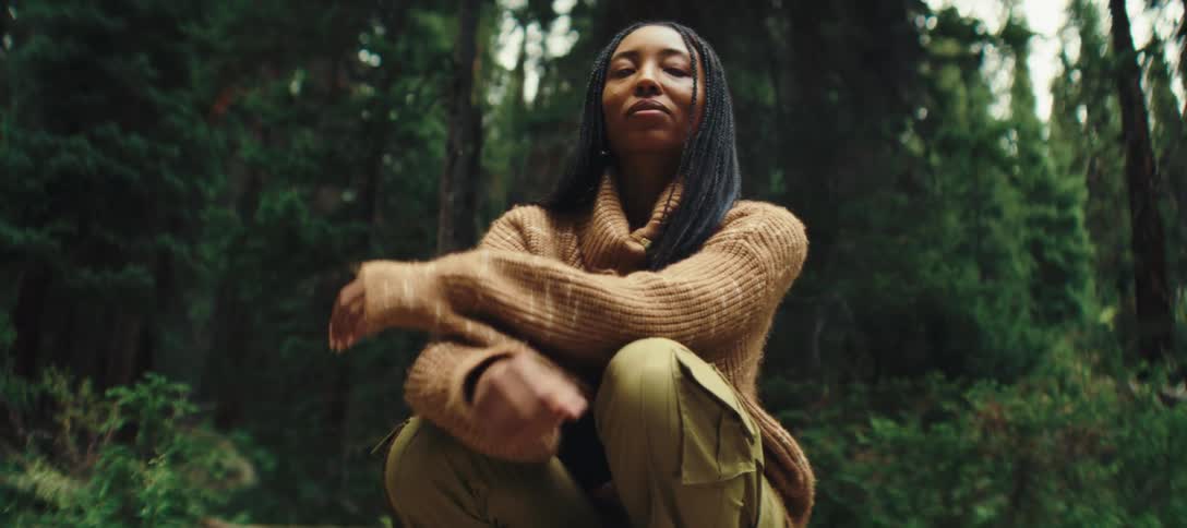 A woman sits crouched in a forest clearing crossing her arms over her chest
