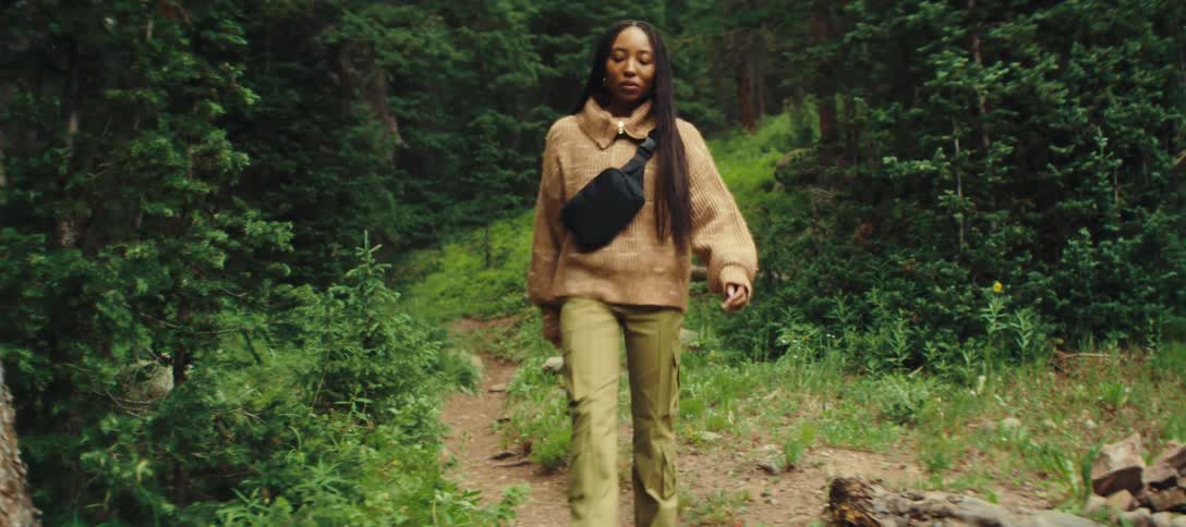 The video shows a young woman with a fanny pack on her chest walking through a forest she looks directly at the camera then turns and walks out of the frame behind a tree