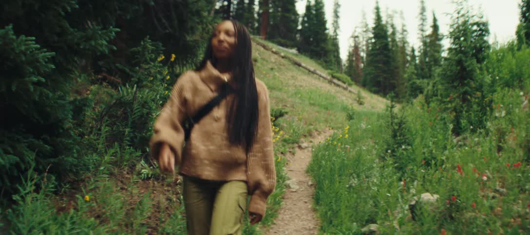 A woman with long dark hair wearing a brown sweater and green pants walks down a narrow dirt path surrounded by tall green grass and scattered flowers on a hillside